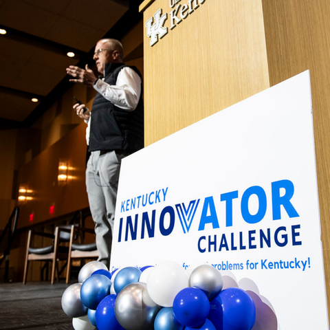 Presenter speaks beside a University of Kentucky podium at the Kentucky Innovator Challenge. Blue, white and silver balloons decorate the stage in a large conference hall.