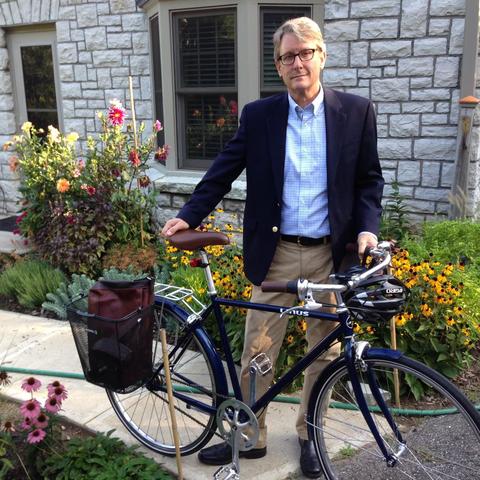 Ned Crankshaw wearing a blazer and blue button down shirt standing on the sidewalk with a bicycle in front of a stone house. 
