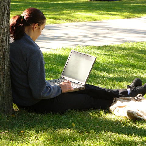 Woman leaning on a tree working on her computer. 
