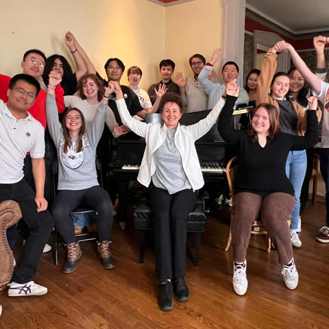 Several college students and a professor gathered around a piano with their hands raised.