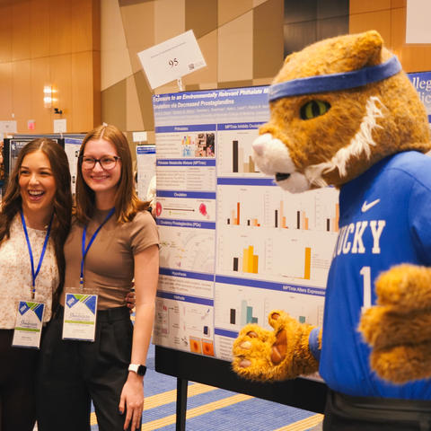 Two students wearing conference badges pose for a photo with the University of Kentucky Wildcat mascot in front of research posters at an indoor showcase event.