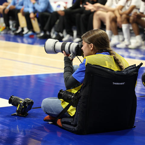 A student with a camera sitting on the floor of a basketball court. 