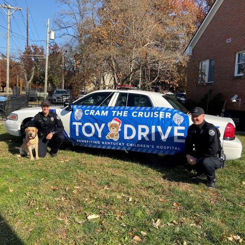 Photo of Officer Michael Culver, K-9 Hudson and Officer Ryan Johnson in front of the cruiser with a banner that says "Cram the Cruiser" toy drive.