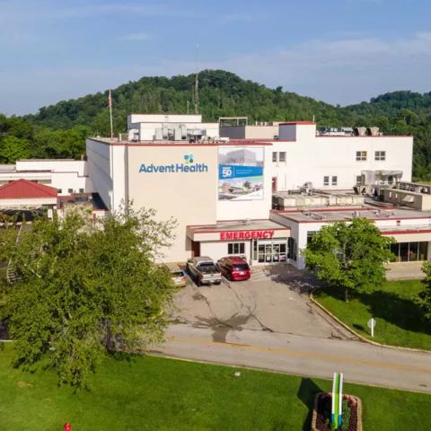 Aerial view of a large light-colored medical building complex with multiple entrances, parked vehicles and ambulance bays, surrounded by green trees and rolling hills under a clear sky.