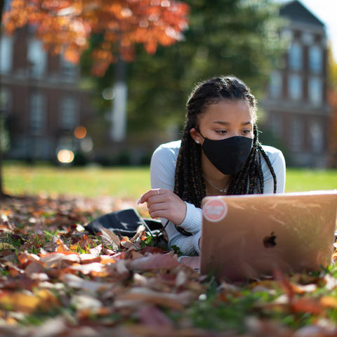 female student wearing mask lying in fallen leaves looking at her computer in front of the Main Building.