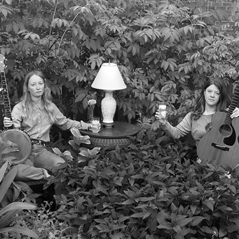 black and white photo of the duo The Other Years seated with banjo and guitar by side table with lamp in overgrown field