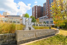 Exterior view of UK HealthCare’s Albert B. Chandler Hospital with its sign in the foreground and surrounding buildings under a bright sky.