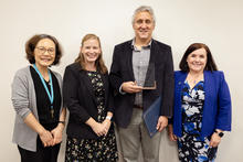 Four people in a group photo, with one person holding a glass award and a blue certificate folder against a plain indoor background.