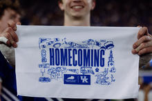 Close-up of a person holding a white rally towel featuring a blue “Homecoming” design with University of Kentucky branding and illustrated campus-themed icons. A blurred crowd and another person are visible in the background.