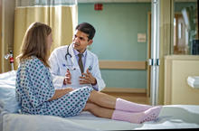 Healthcare provider sits beside a patient on a hospital bed, speaking and gesturing during a one-on-one conversation in a clinical exam room.