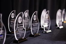 Row of clear glass award plaques displayed on a black table, with the nearest trophies in focus and others blurred in the background.