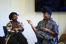 Two Black women seated next to each other in a classroom. The woman on the right is speaking and gesturing, and the woman on the left is listening.