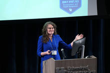 Speaker stands at a podium at the University of Kentucky, gesturing while presenting slides. A microphone and monitor are on the podium, and a projected screen behind displays the John P. Wyatt, M.D. Environment and Health Symposium logo.