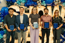 Five individuals stand side by side holding framed awards in front of a balloon arch backdrop.
