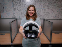 Person standing between office cubicles holds a virtual reality headset toward the camera. A wall-sized whiteboard covered with colorful sketches, diagrams and notes fills the background of the workspace.