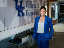 A person in a blue suit stands in a hallway beside a wall displaying the University of Kentucky logo and the words “College of Education.” A counter with stools lines the wall, and the hallway extends into the background.