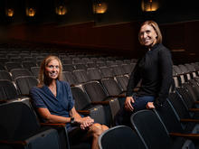 Two people pose among rows of seats in a dimly lit auditorium, with one seated and the other leaning against a nearby chair. Wall lights illuminate the theater in the background.