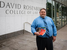 A person stands outside the Rosenberg College of Law building, holding several books.