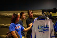 Three students browse a clothing rack, with a white “Kentucky Wildcats” shirt displayed in the foreground.
