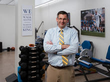 Person stands with arms crossed in a biomechanics lab containing dumbbells, exercise equipment and wall displays, including a sign reading, “We cannot walk alone.”