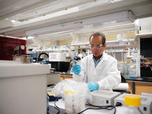 A researcher wearing a white lab coat, safety glasses and blue gloves uses a pipette while working at a laboratory bench surrounded by scientific instruments, bottles and testing equipment in a University of Kentucky research lab.
