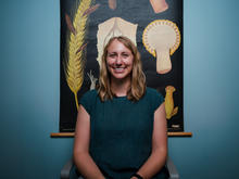 Person seated in a chair poses in front of a botanical wall chart featuring enlarged illustrations of fungal structures on a dark background.