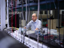A researcher sits at a computer workstation in a laboratory, viewed through an array of cables, instruments and scientific equipment. 