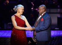 Two people in formal attire hold hands on a lit stage, one in a red sequined gown and the other in a dark suit with a red pocket square.