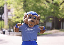 A University of Kentucky Wildcat mascot stands outdoors wearing a blue “One University at Kentucky” T-shirt.