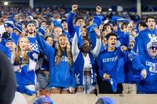 A crowd dressed in blue University of Kentucky apparel raises arms and cheers at a campus event.