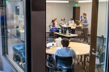 A view through a glass window into a classroom or meeting room where several people are seated around tables, engaged in discussion or work.