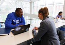 Two people sit across from each other at a table in a campus study space, with one using a laptop displaying UK-related stickers and the other engaged in conversation.
