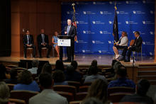 A man speaks at a podium before a UK Markey Cancer Center backdrop, with five people seated onstage and an audience in the foreground.