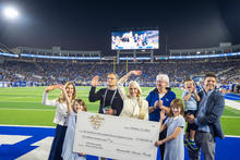 Group of adults and children standing on a football field at night, holding a large ceremonial check, with a packed stadium and scoreboard in the background.