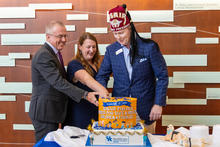 Three people are cutting a decorated cake on a table. The cake reads “Kosair for Kids – Hope for Safe & Healthy Children & Families” and features blue and yellow colors.