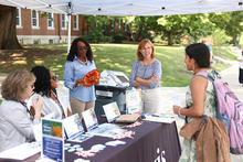 People speak with visitors at an outdoor information table under a tent on campus, with brochures, handouts and a landfill waste display.