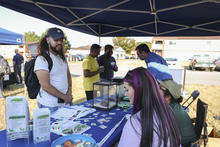 A community outreach event taking place under a blue canopy tent. A table covered with a blue tablecloth displays brochures, stickers, and small containers, while two seated participants interact with a standing attendee wearing a backpack. 