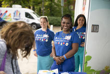 Three people wearing blue “Be Healthy Bash” shirts stand beside a mobile health unit and greet an attendee at an outdoor event. A table with event materials is in the foreground, and a branded vehicle is visible in the background.