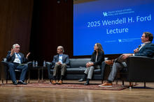 Four panelists sit on a stage during a public lecture and discussion at the University of Kentucky. One speaker gestures while talking from an armchair, while the other panelists listen from a sofa and adjacent chairs. A large screen behind them.
