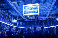 Graduates in caps and gowns stand inside Rupp Arena as blue streamers fall from the ceiling, with a large scoreboard displaying the celebration overhead.