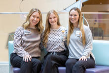 Three people seated closely together on a cushioned bench in an indoor setting, each wearing long-sleeved patterned tops and dark pants