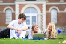 Three students sitting on grass outside a campus building, smiling and looking at a laptop together.