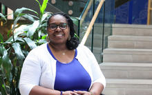 Person seated indoors near a large leafy plant and a staircase, wearing glasses, a blue top and a white cardigan, with hands folded and facing the camera.