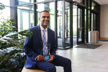 A person in a blue suit and University of Kentucky-patterned tie sits on a bench in a bright lobby, holding a glass award.