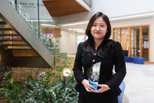 Person in a black outfit standing in a modern building lobby and holding a clear glass award with a blue base. A staircase with glass railings, indoor plants, and seating areas are visible in the background. 
