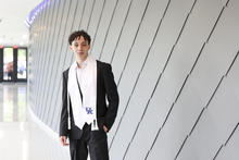 A student stands in a modern campus building hallway, next to a curved wall with geometric paneling.