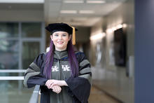 A University of Kentucky graduate wearing doctoral regalia and a black academic cap with a gold tassel stands smiling in a hallway. The graduate’s purple hair and UK logos on the gown are visible as they rest their arms on a railing indoors..