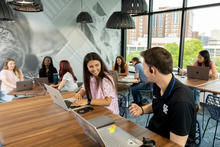 Students work and talk together around tables with laptops in a bright campus study space.