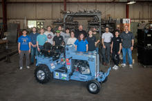 A group of about 15 people stand in a workshop or fabrication space behind a small, blue custom-built tractor labeled “Wildcat 3526 22.” The tractor, covered with sponsor logos, sits in the foreground.