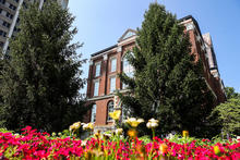 Historic brick building framed by flowers and evergreen trees.